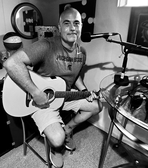 Black and white image of a bald white man with a guitar, sitting on a stool.