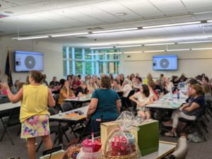 Image of meeting room with tables filled with attendees, tv displays on back walls, and a foreground table with gifts and prizes.