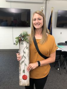 Blonde Caucasian woman in yellow shirt holds rustic painted board with pine bough and red ornament motif.