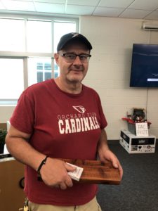 Man in red t-shirt and baseball cap holds cribbage board and deck of cards