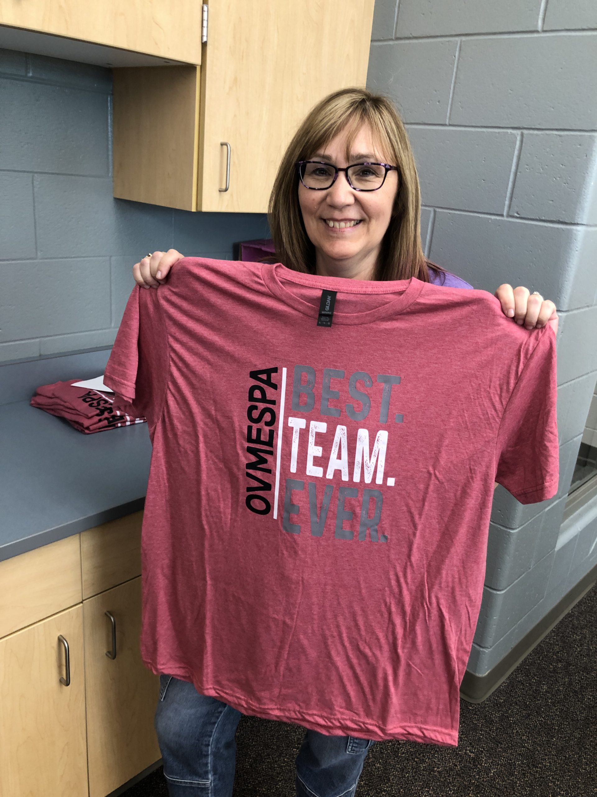 Caucasian woman with dark-framed glasses holds up a red t-shirt that reads "OVMEPSA | BEST. TEAM. EVER."